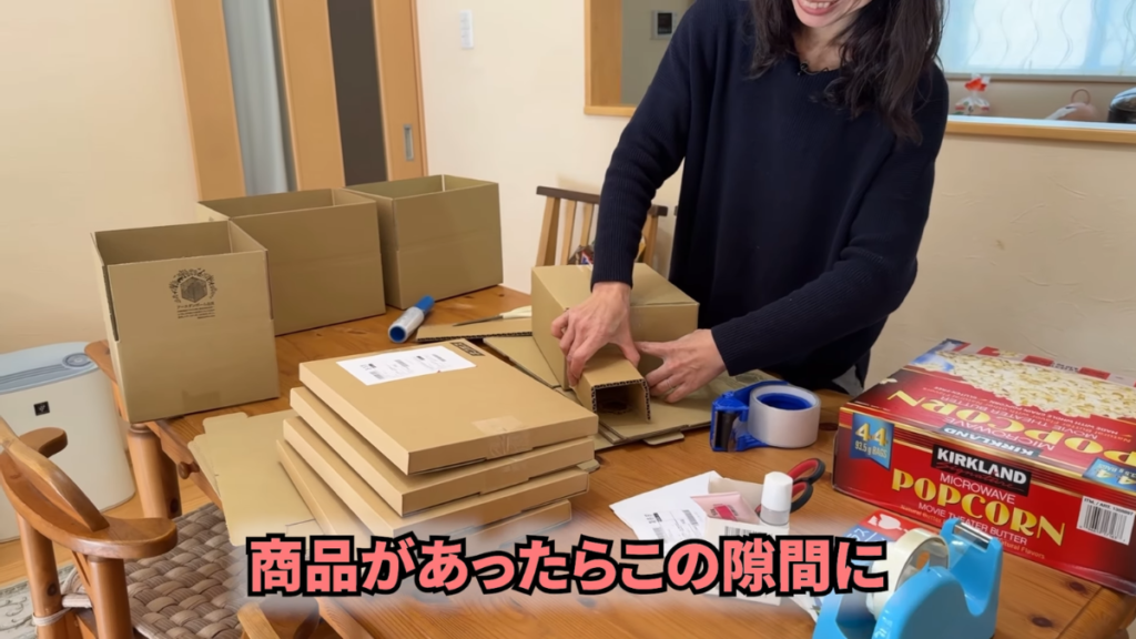 リビングのテーブルで商品の梱包作業をしている。緩衝材を購入する代わりに、不要になった小さな段ボールを折りたたんで商品の隙間に詰め込み、コスト削減と商品保護を両立させている様子。